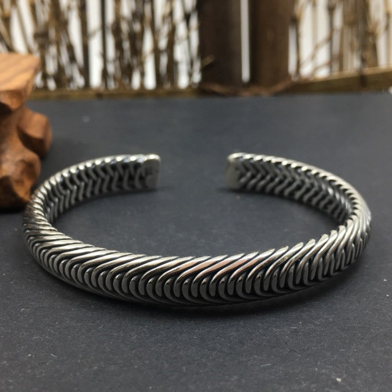 The heavy Nordic Frost Braided Silver Cuff resting flat on a dark slate surface with a soft-focus wooden accent in the background. The low-angle lighting accentuates the mirror-polished metallic finish and the rugged herringbone texture of the 35g solid silver band.