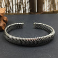 The heavy Nordic Frost Braided Silver Cuff resting flat on a dark slate surface with a soft-focus wooden accent in the background. The low-angle lighting accentuates the mirror-polished metallic finish and the rugged herringbone texture of the 35g solid silver band.