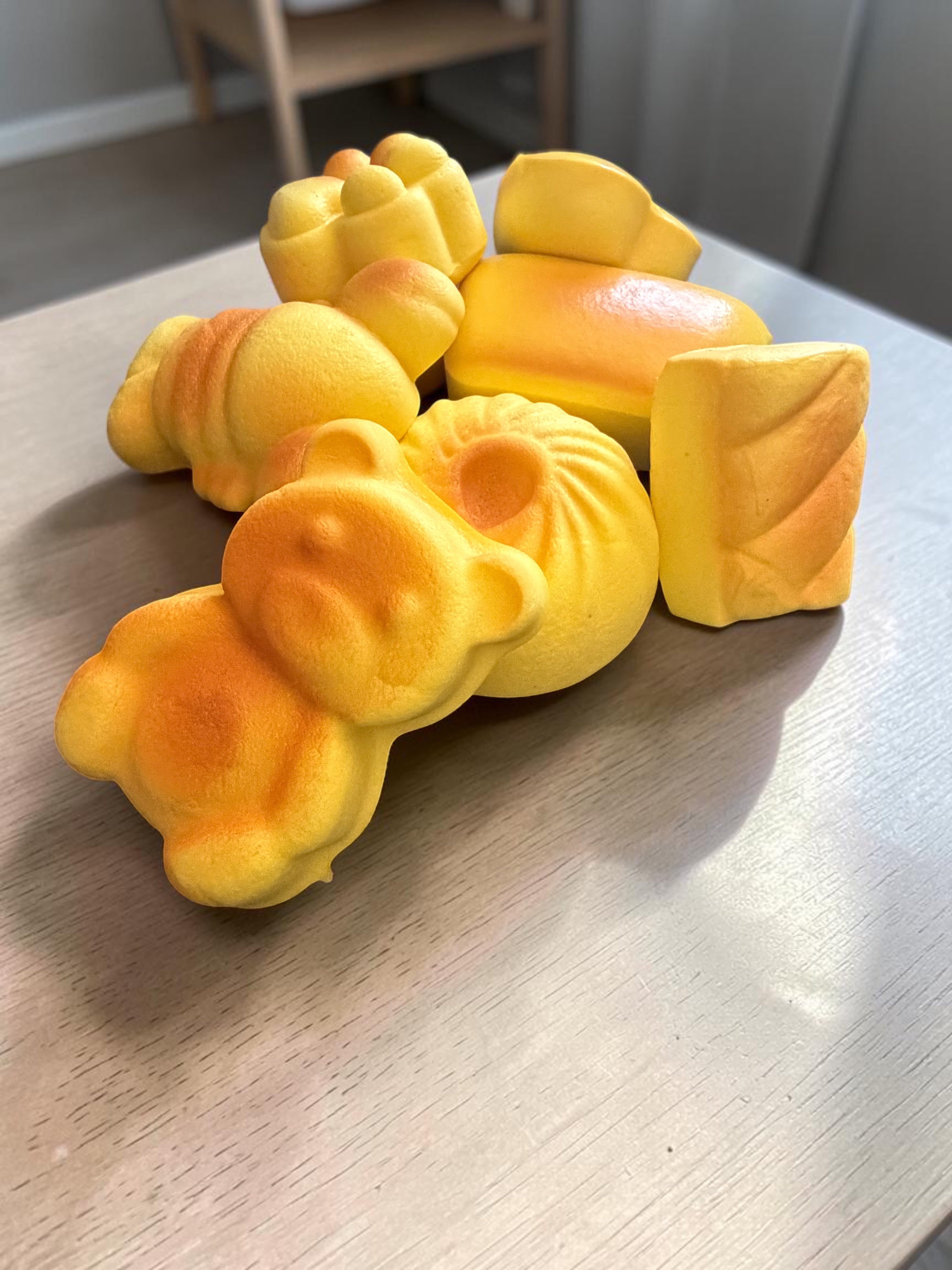 A group of yellow novelty bread-shaped items, including a bear-shaped piece, a croissant, a bun, and a rectangular loaf piece, arranged on a light-colored wooden tabletop.